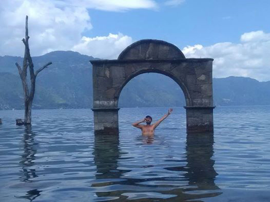A swimmer standing in the water inside a stone arch that stands in Lake Atitlán, with mountains behind